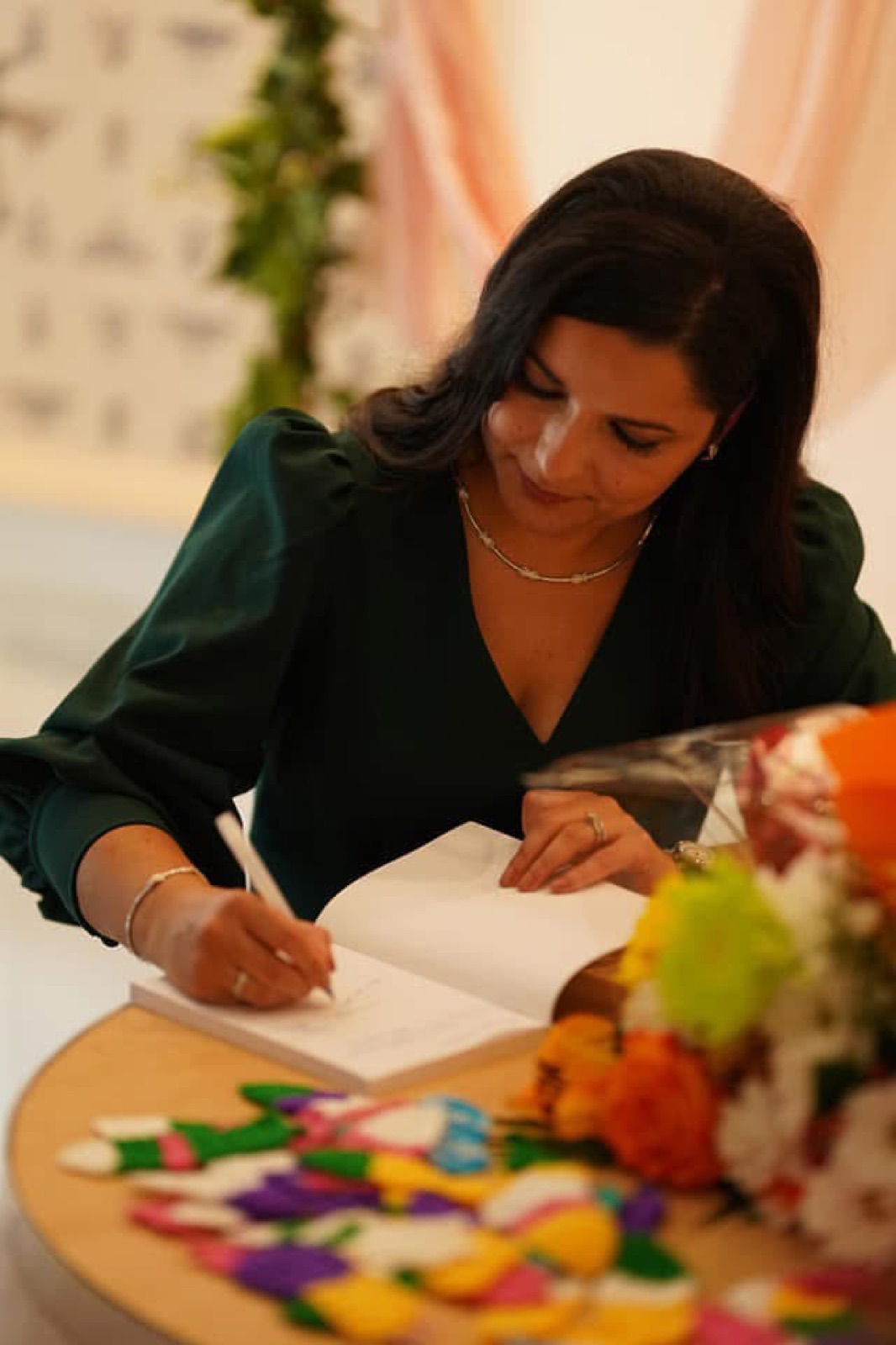 Gros plan de Salam Chaya en robe verte signant un exemplaire de Mourir d'aimer, table ornée de confettis colorés et de fleurs