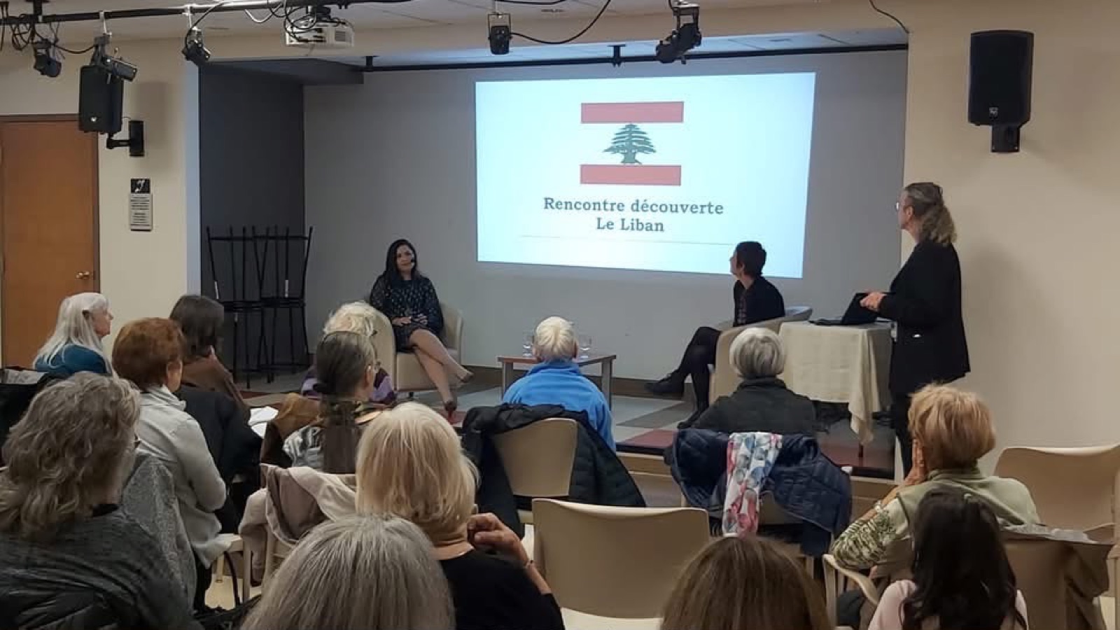 Salam Chaya assise sur scène, drapeau libanais projeté en arrière-plan, devant un auditoire attentif